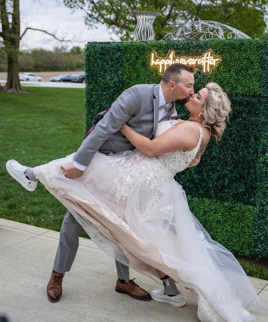Couple in wedding attire embracing in front of a green hedge with 'Happy Anniversary' sign.